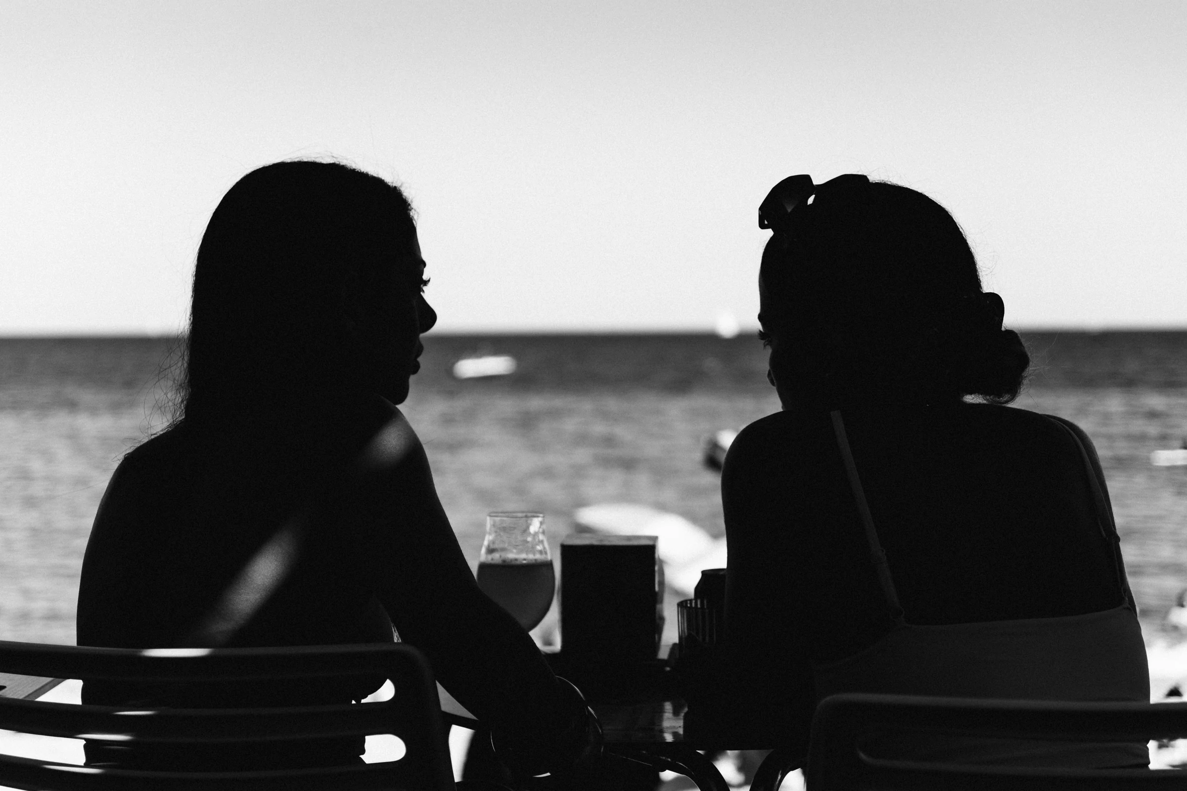 Two women in silhouette at a café table, the sea behind them