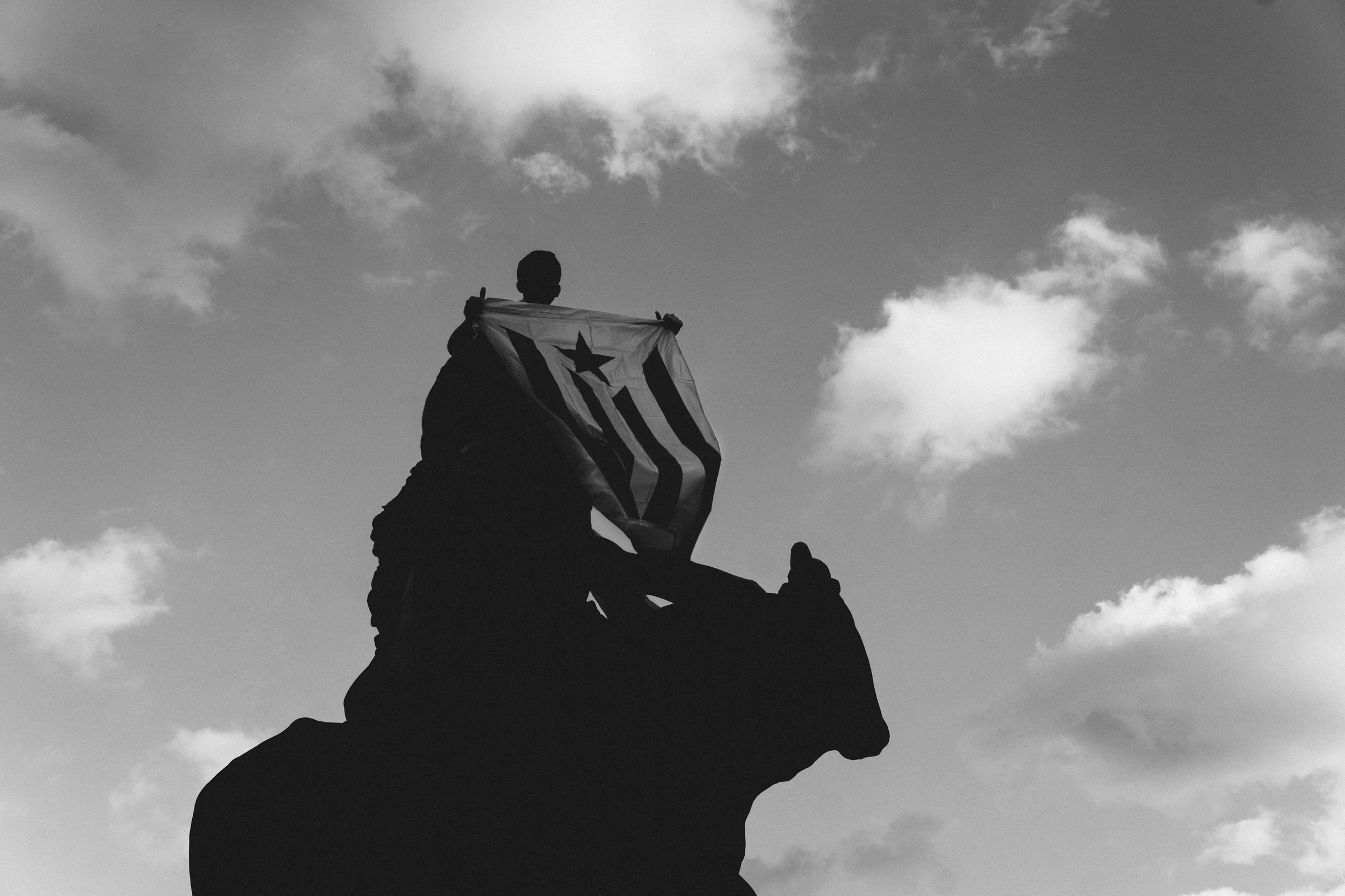 A boy holding up a striped flag on top of a statue, in silhouette against the clouds