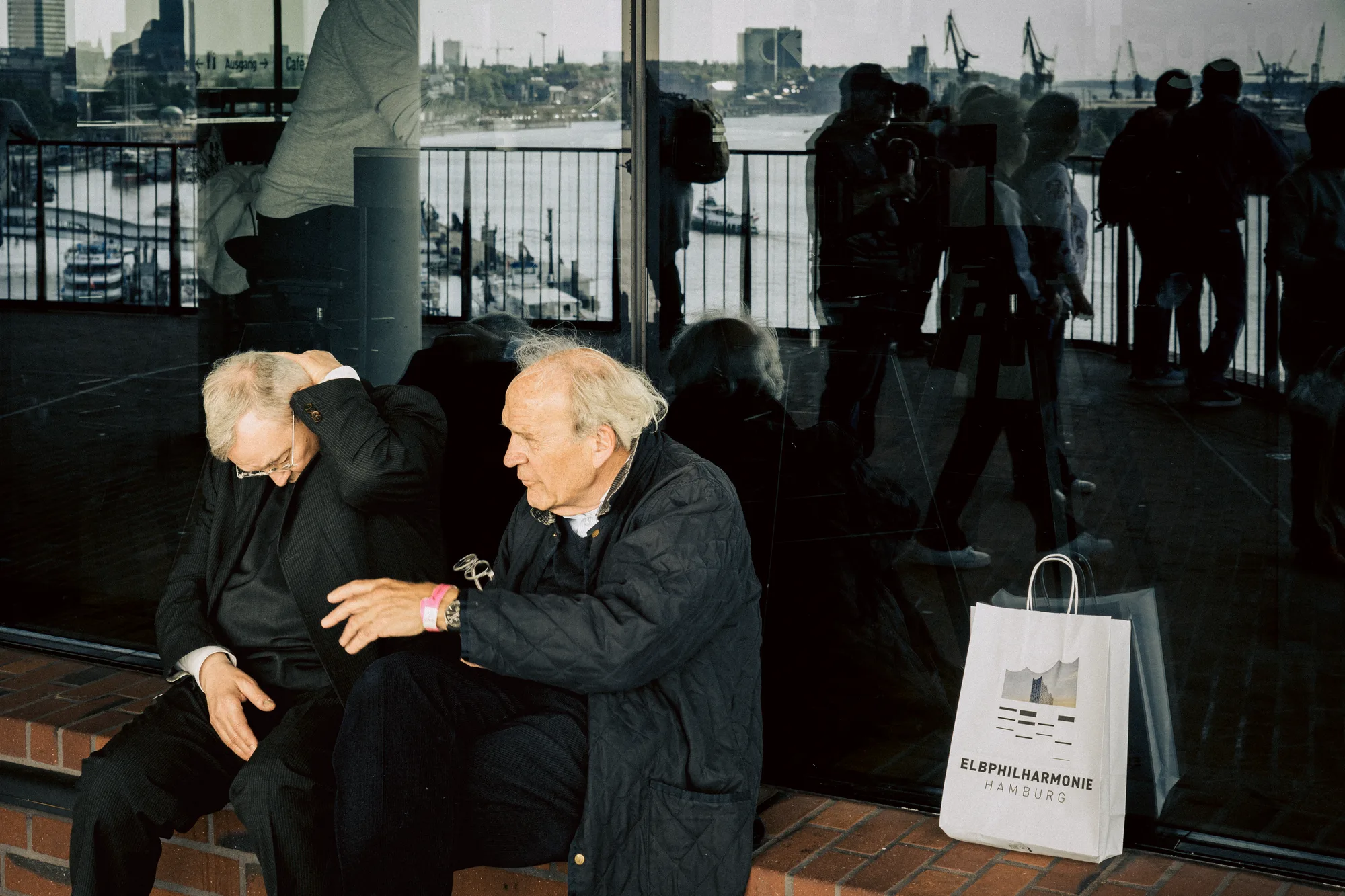 Two older men resting on a ledge at the Elbphilharmonie, the harbour reflected behind them