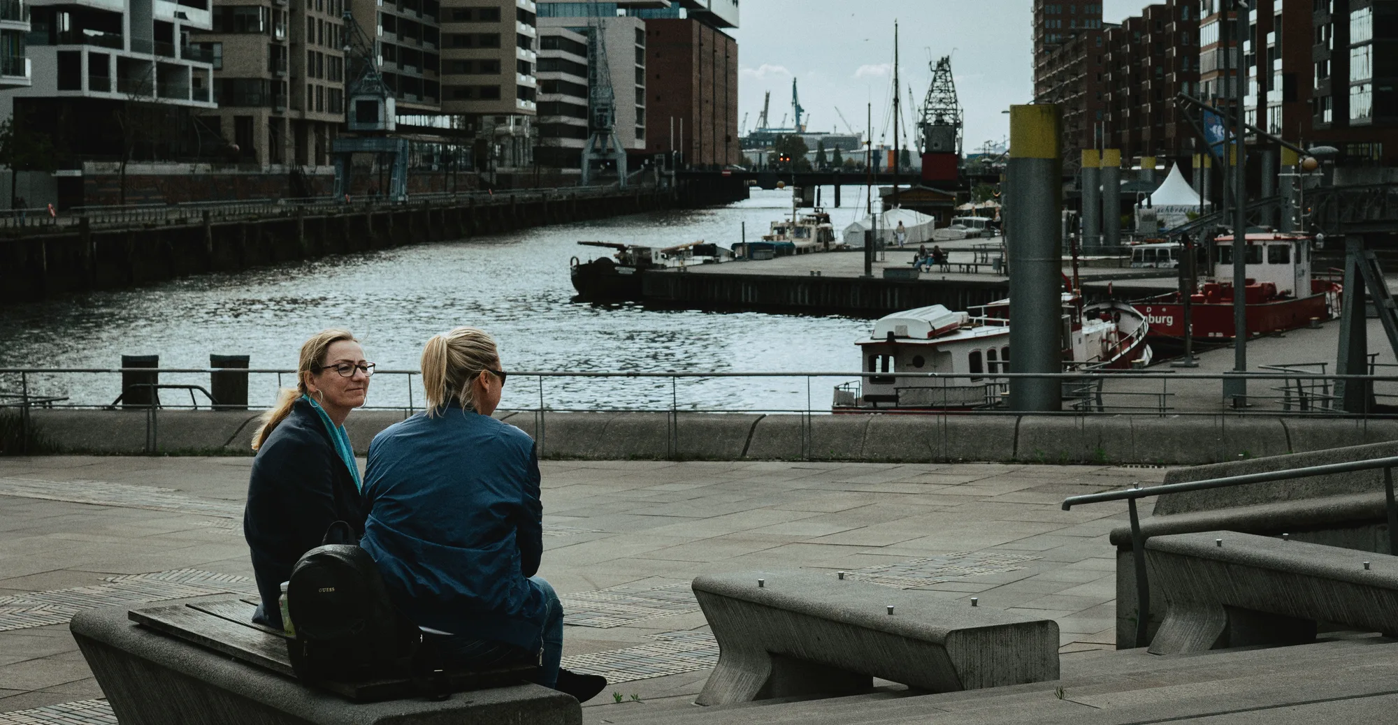 Two women talking on a bench by the harbour, boats and cranes behind them
