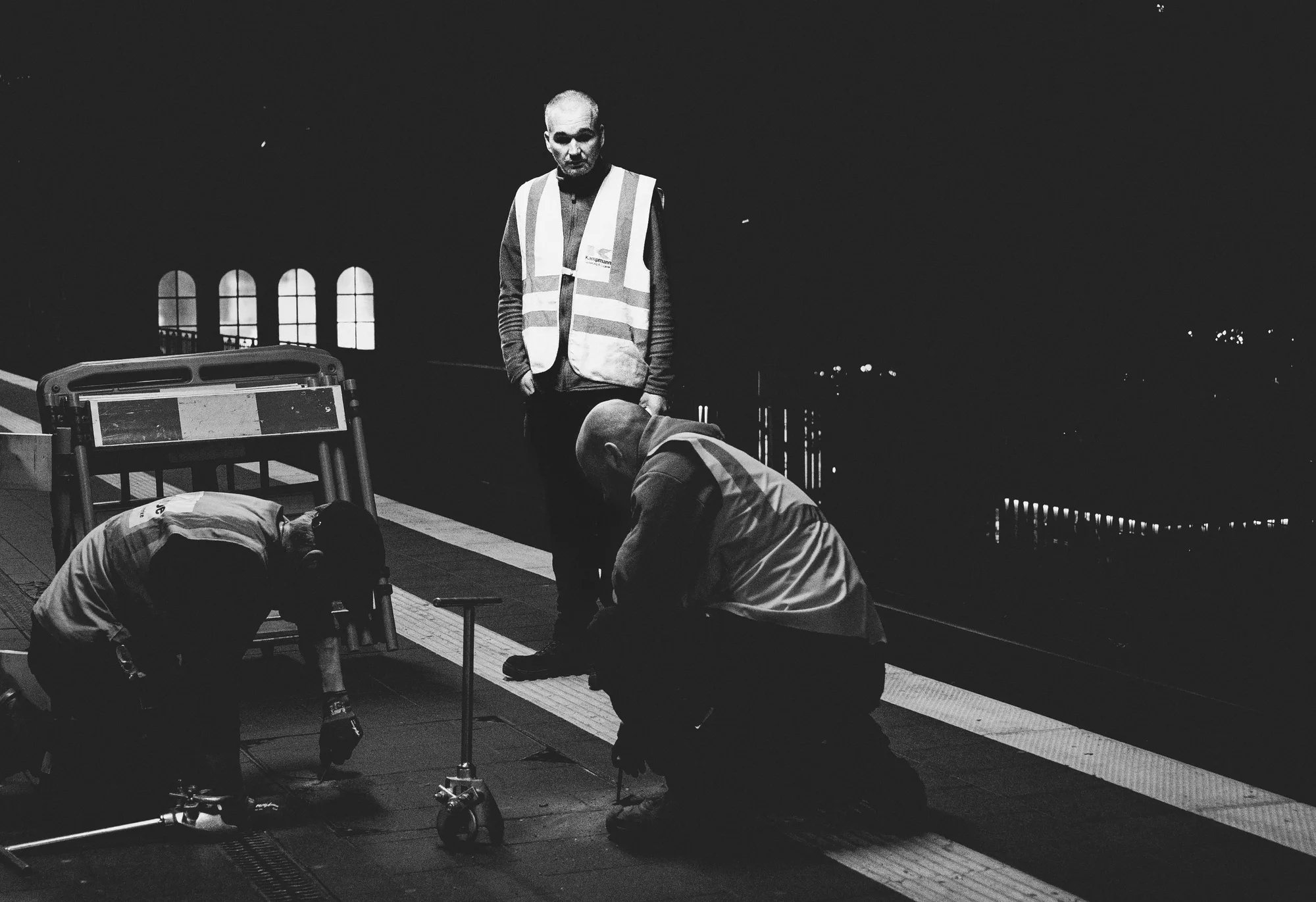 Two workers kneeling on a station platform while a third stands watching