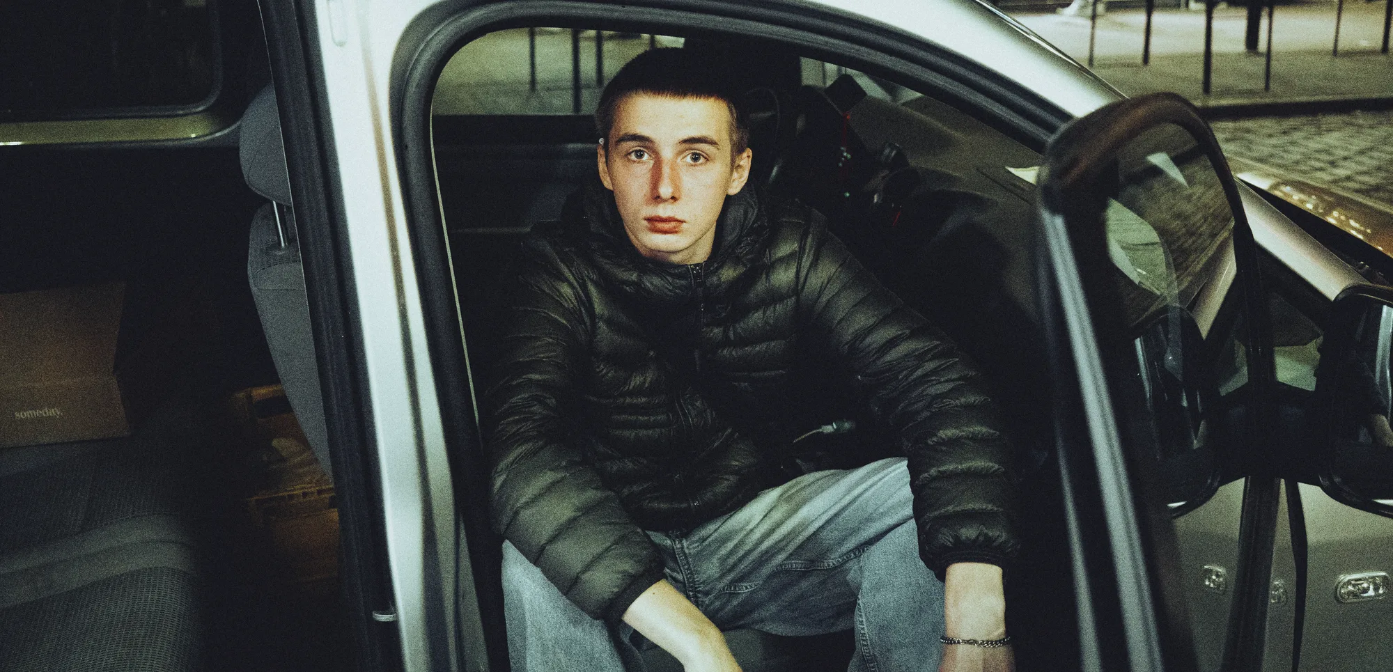 A young man sitting in the open door of a car, looking at the camera