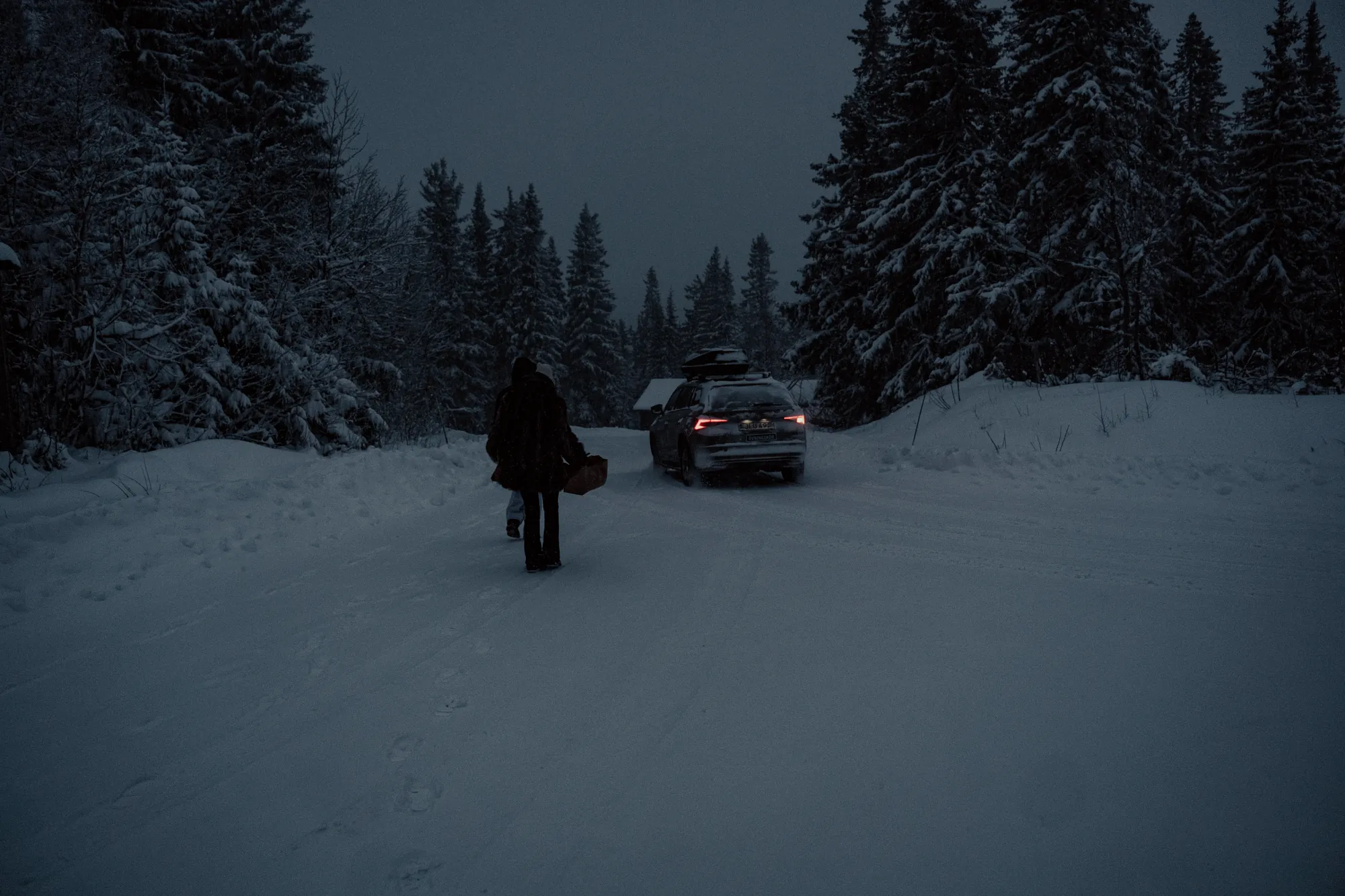 A figure walking towards a parked car on a snowy road at dusk