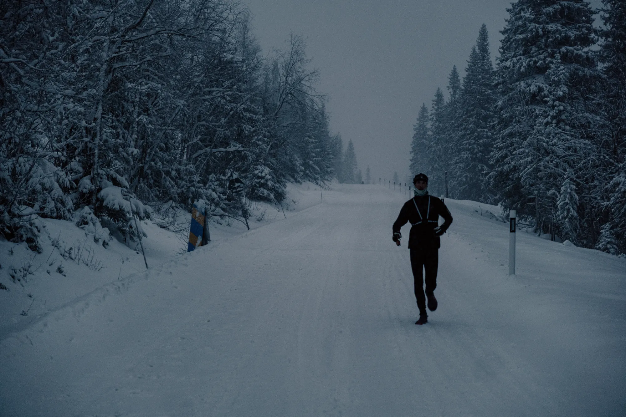 A runner in a reflective vest coming down a snowy road through the forest