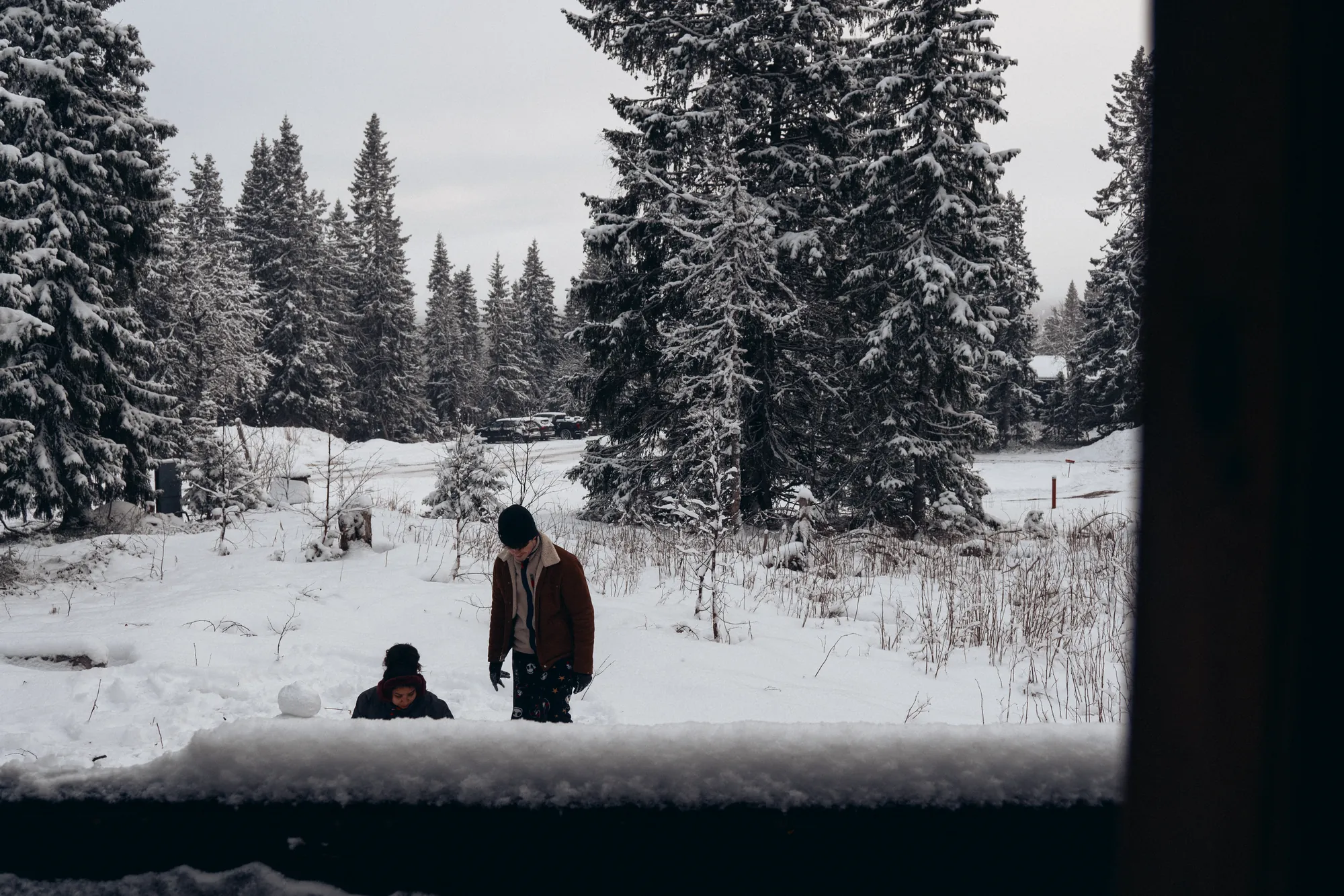 Two people building a snowman, seen through a cabin window with snow on the sill