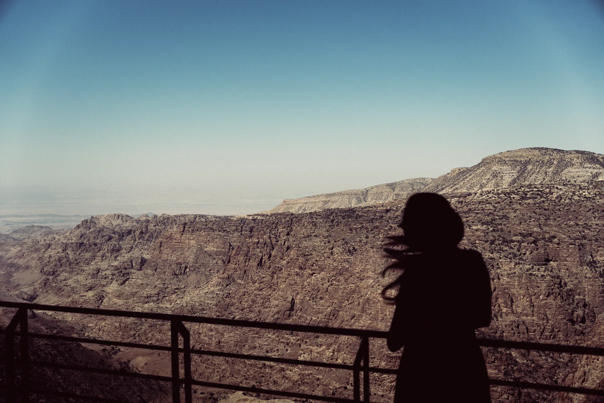 A woman in silhouette at a balcony railing above the Dana valley, her hair in the wind