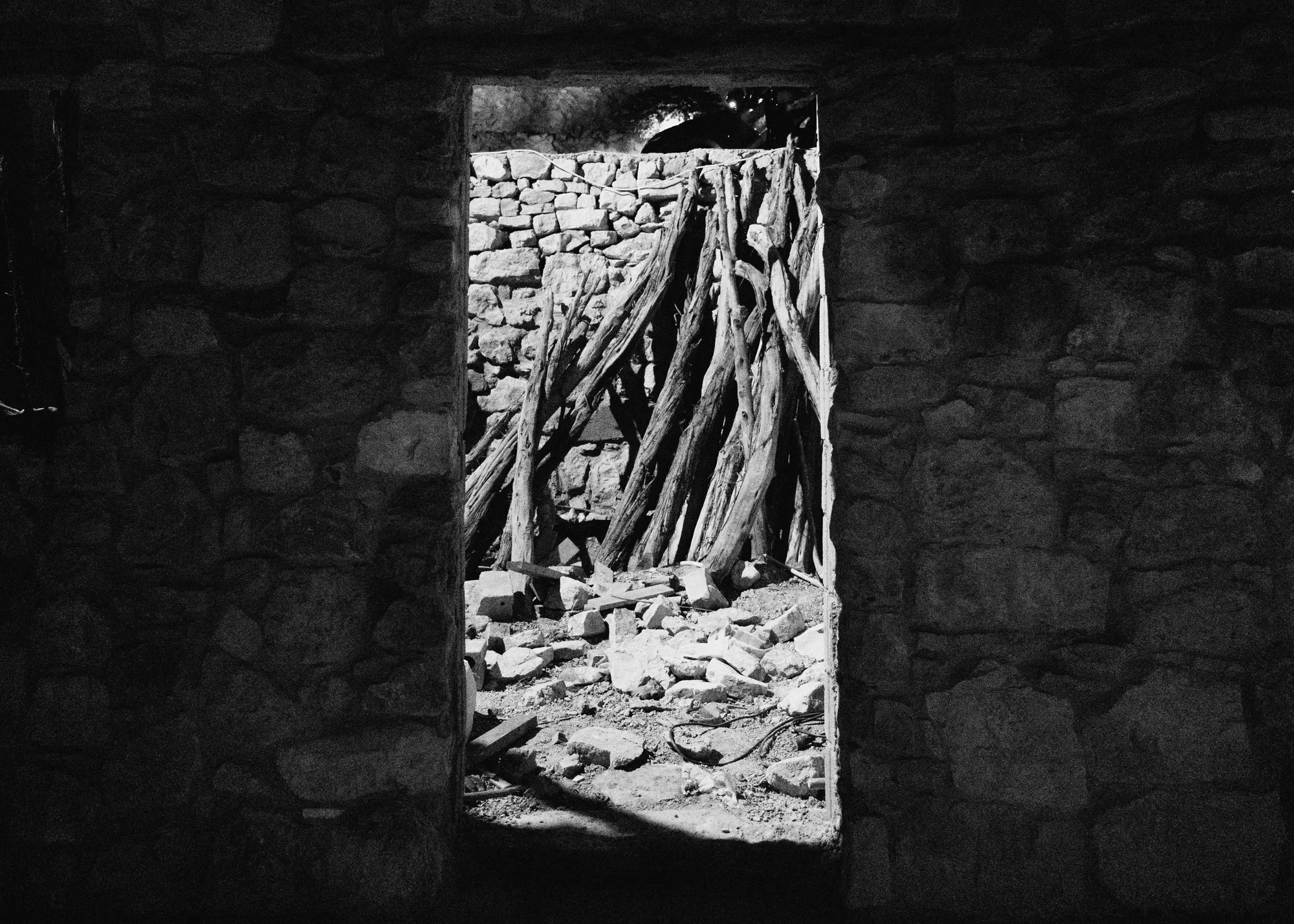 A doorway in a dark stone wall opening onto a pile of cut branches and rubble