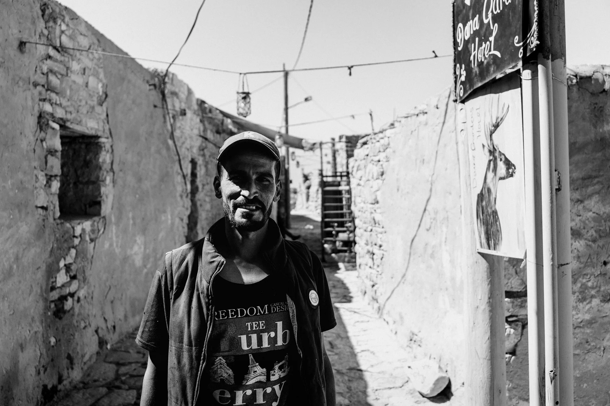 A man in a cap standing in a narrow stone lane beside a hand-painted hotel sign in Dana