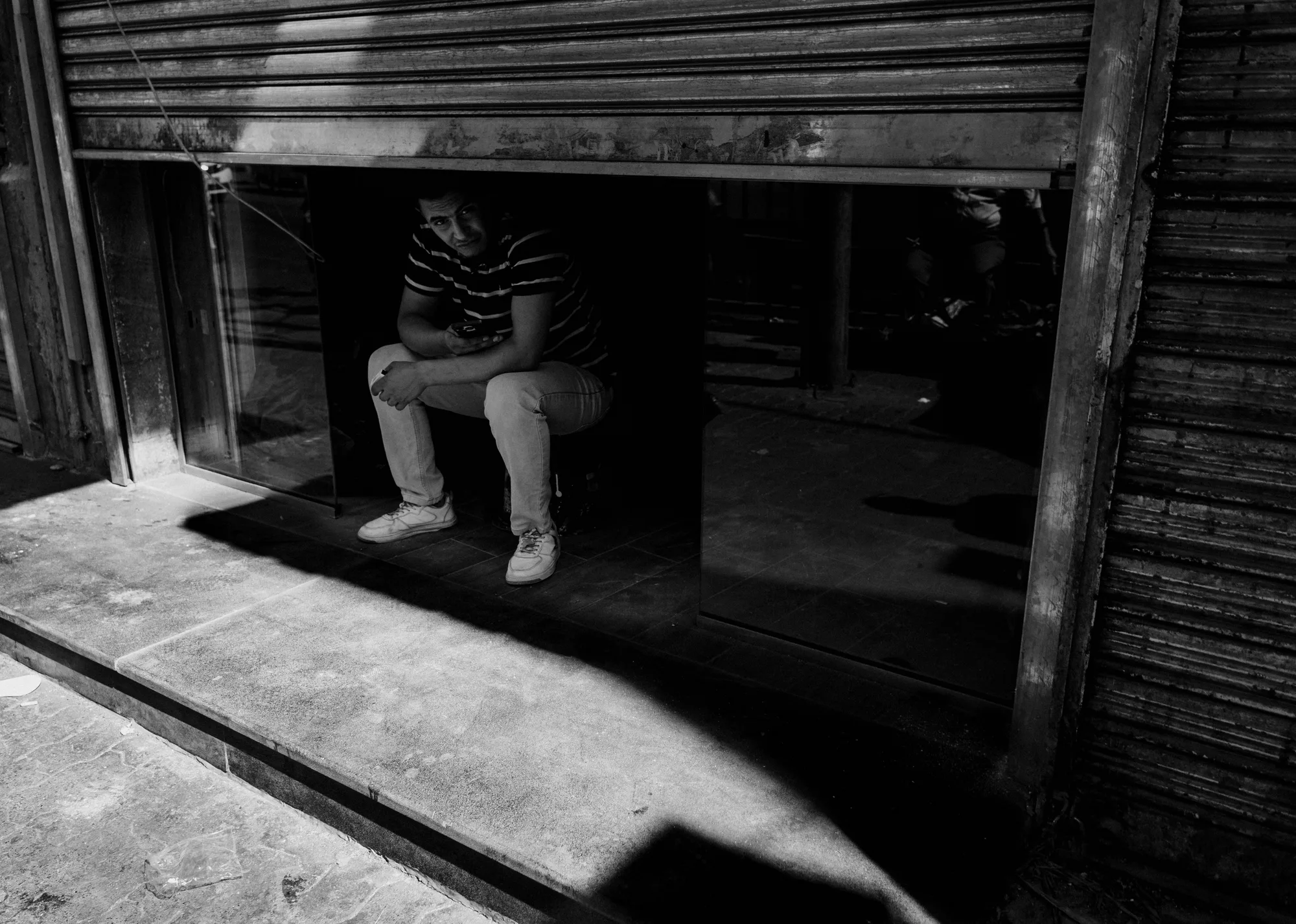 A man sitting in the dark of a half-open shopfront, looking out at the bright street