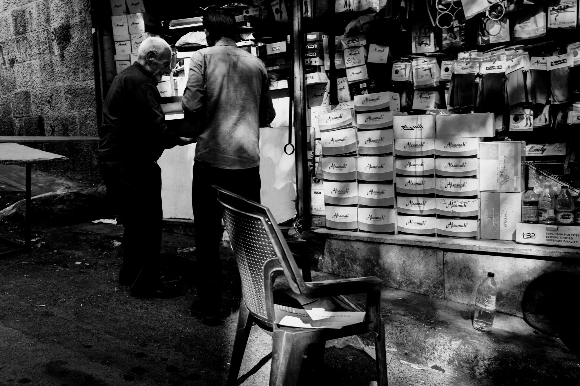 An old man and a younger man at a stall stacked with sock boxes, an empty plastic chair in front