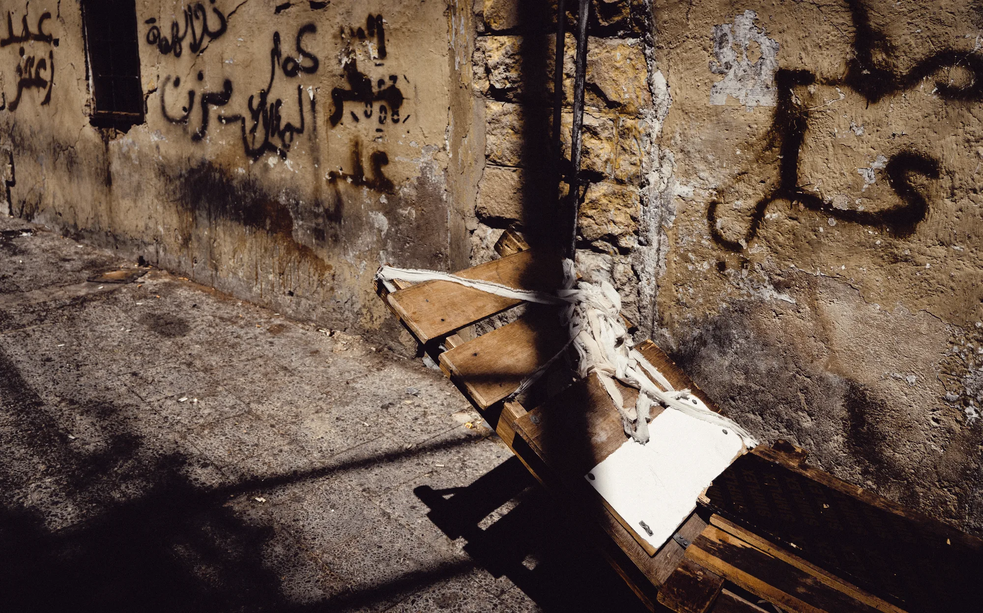 A broken bench tied to a pole in a sunlit alley with Arabic graffiti on the wall