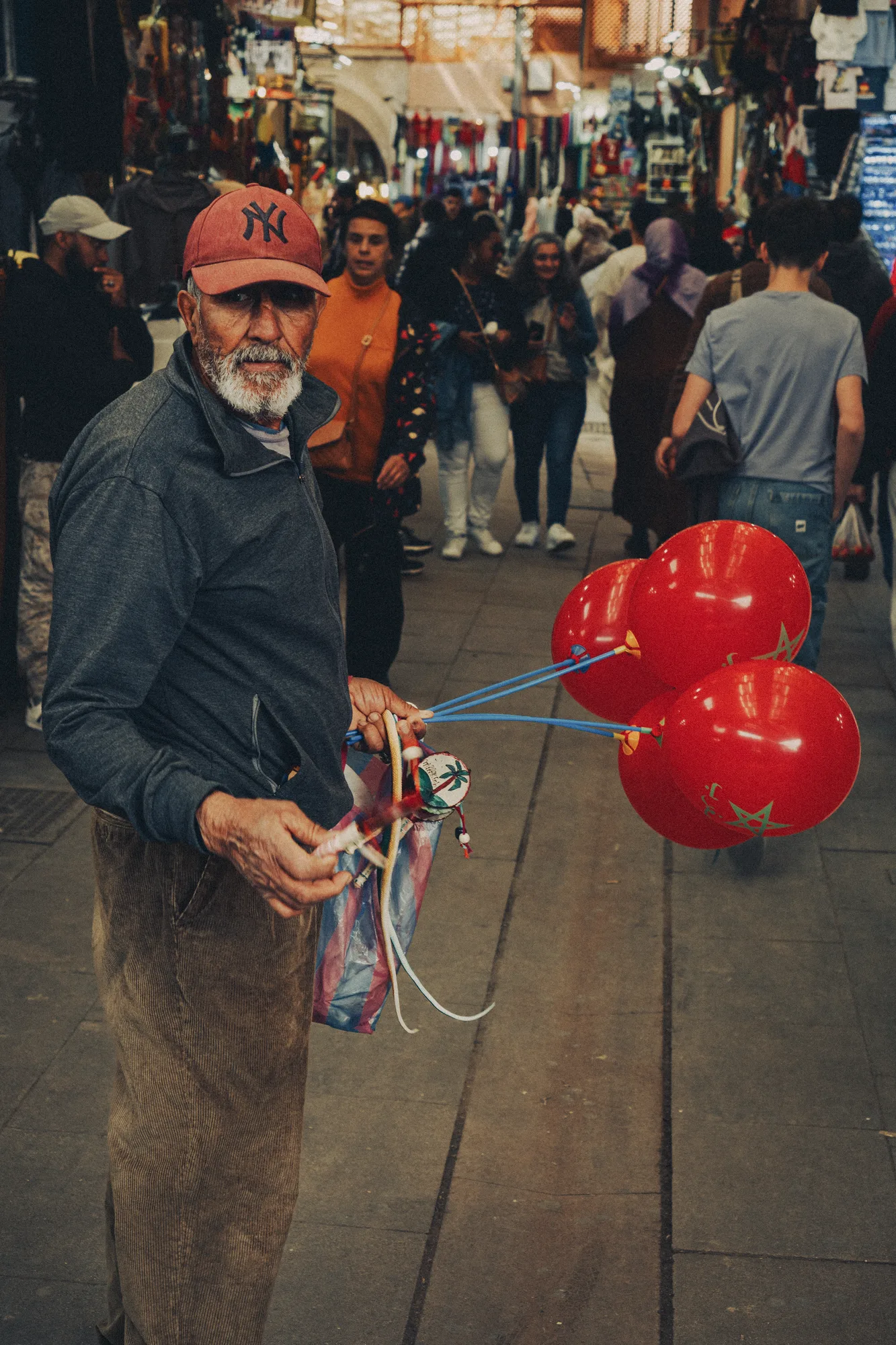 An older man in a red cap holding a bunch of red balloons in a busy souk