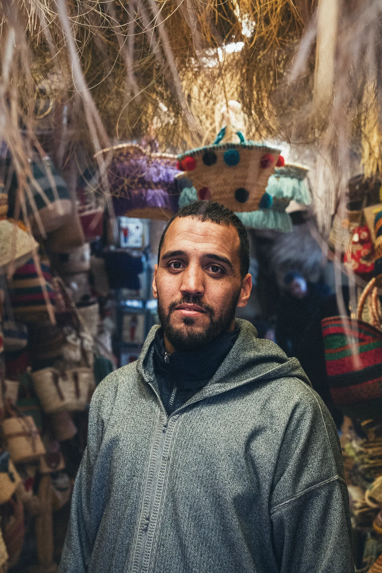 A man in a grey djellaba standing under hanging baskets and bags