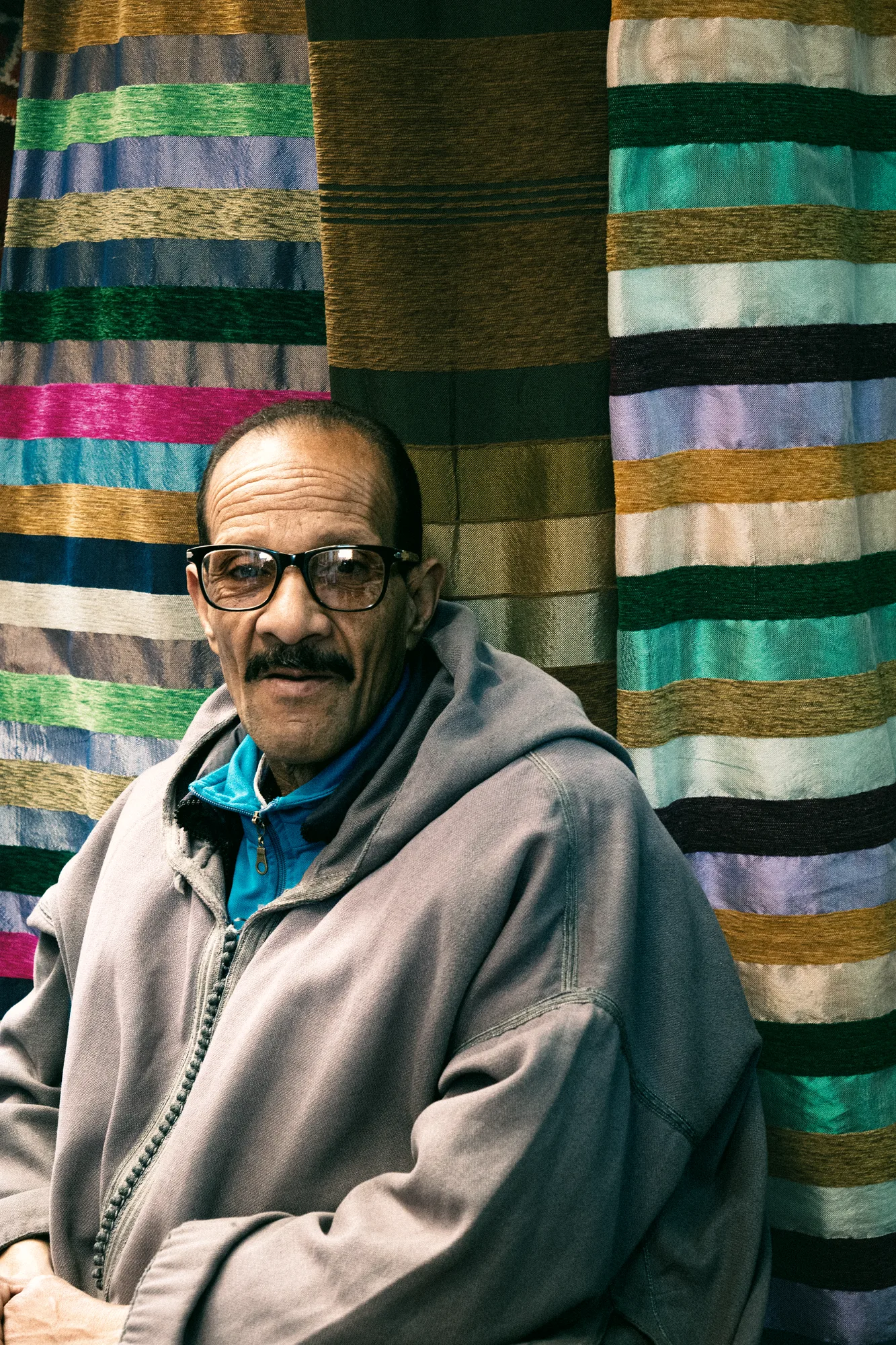 A man in glasses and a djellaba smiling in front of striped woven rugs