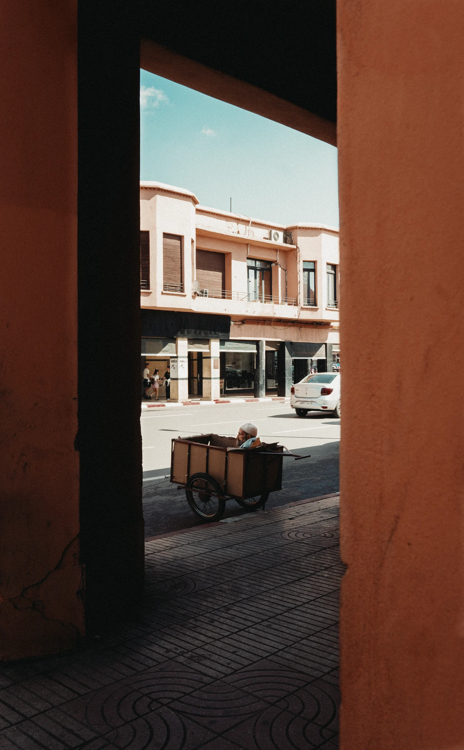 A man pulling a cart along a sunny street, seen between two orange columns
