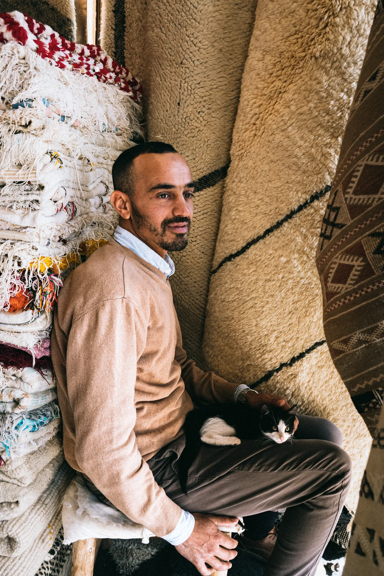 A man leaning against rolled rugs with a black and white cat asleep on his lap