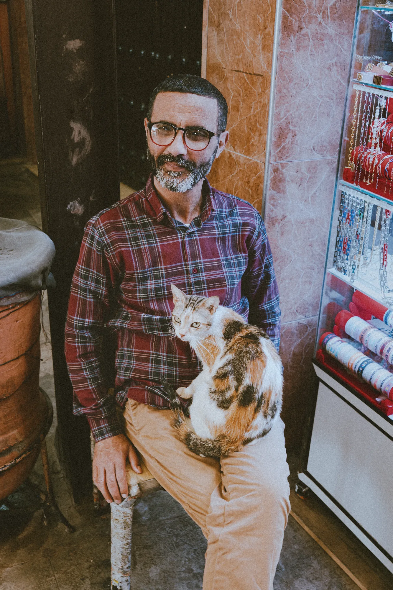A man in a checked shirt sitting in a shop doorway with a cat on his lap