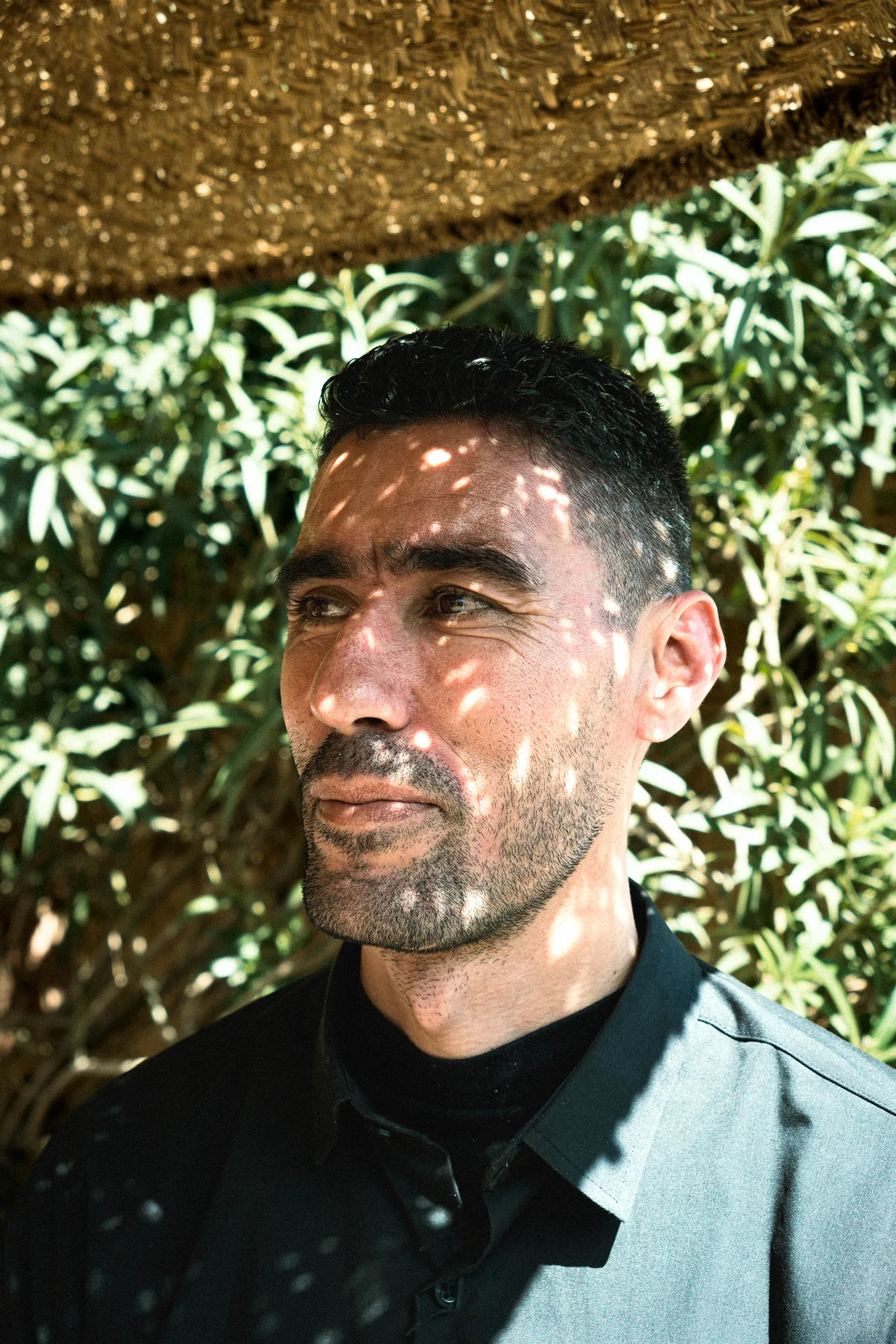 A man in dappled sunlight under a straw awning, looking to the side