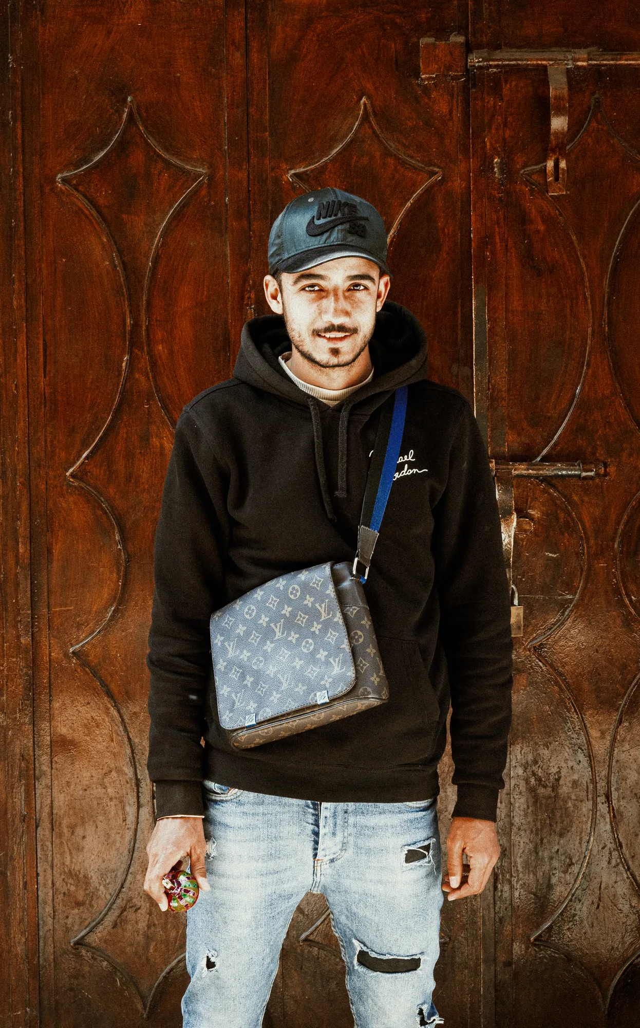 A young man in a cap and hoodie with a patterned shoulder bag in front of a carved wooden door