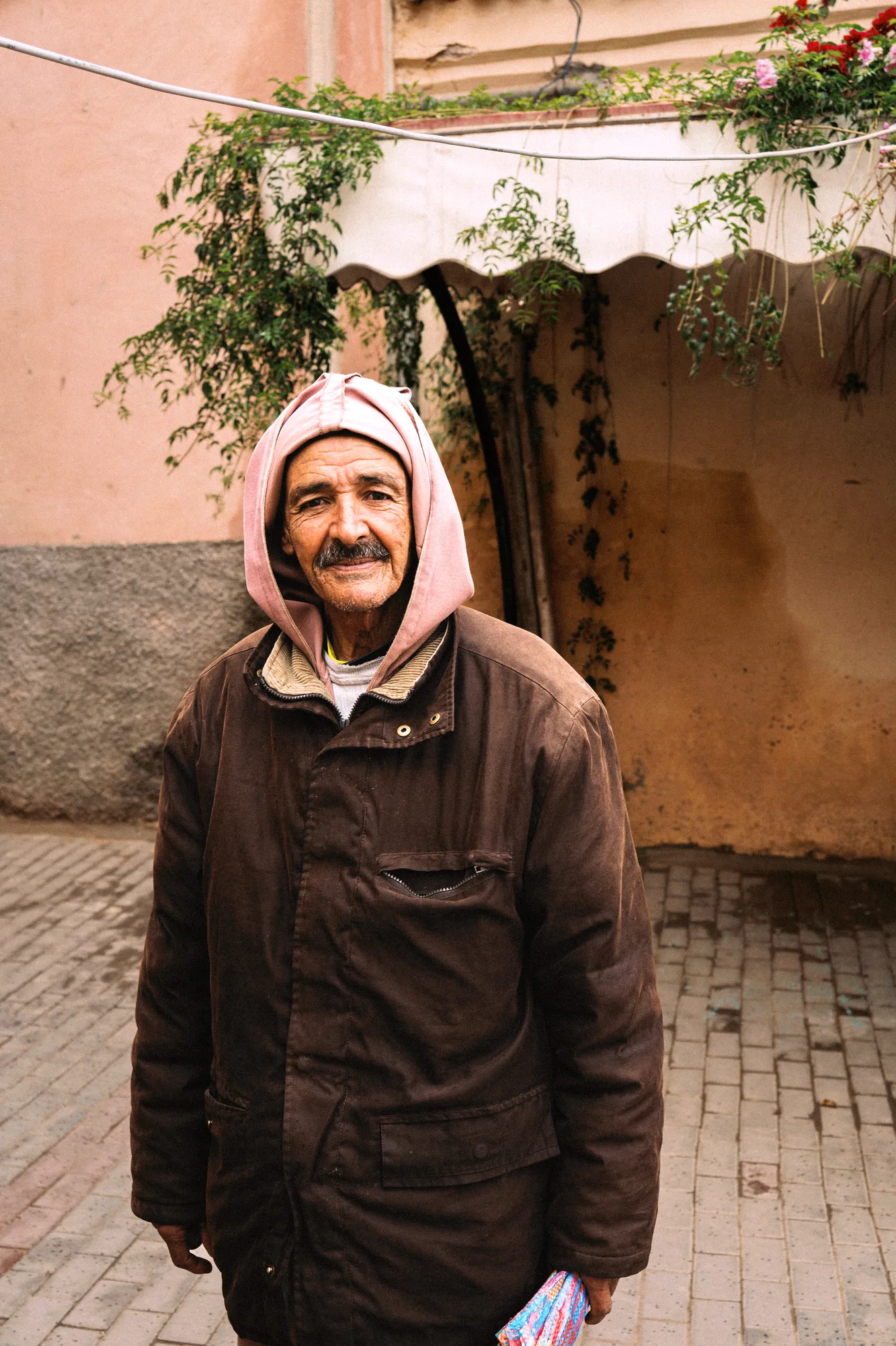 An older man in a pink hood and brown jacket standing in a courtyard