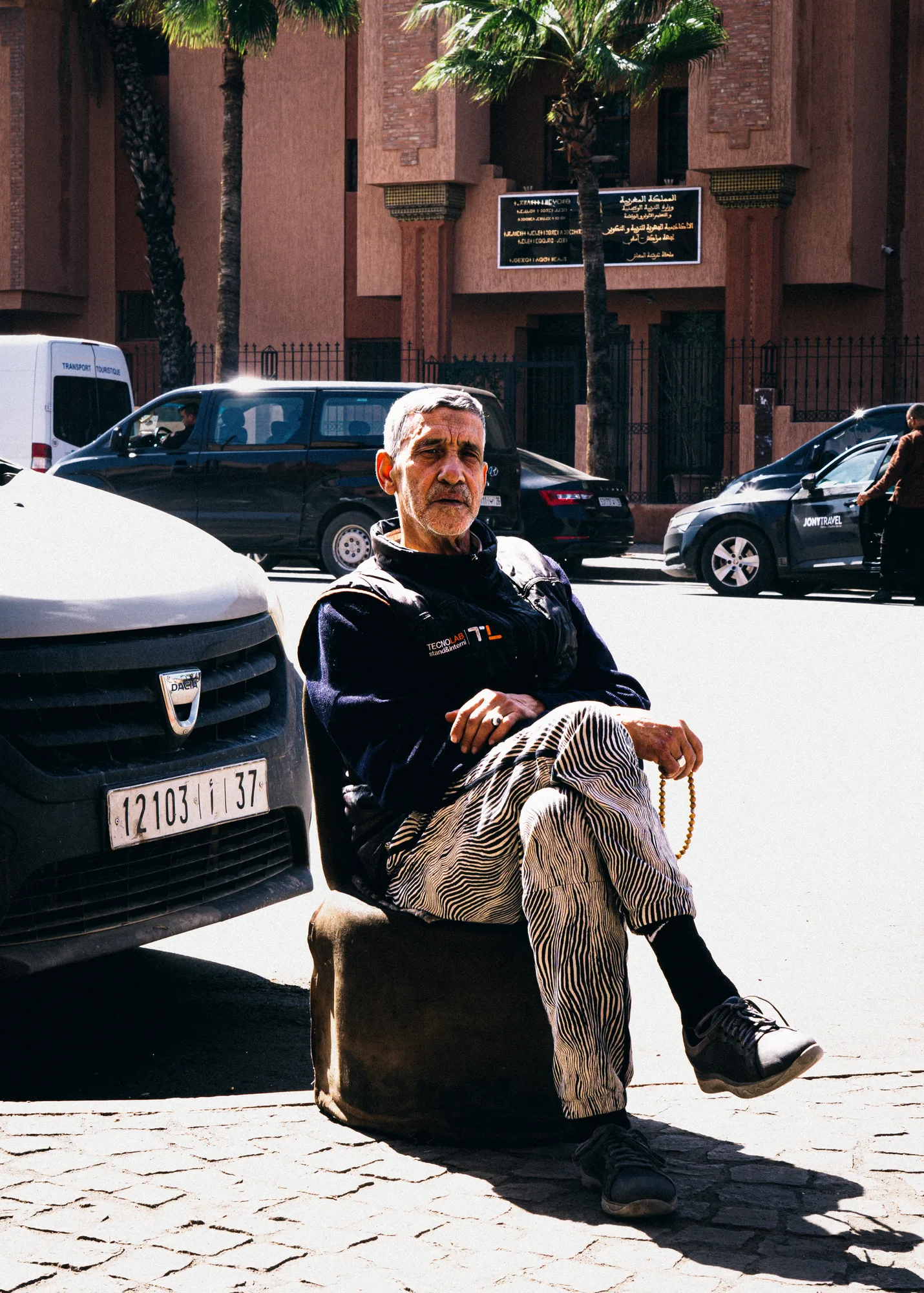 An older man with prayer beads sitting cross-legged on a stool beside a parked car