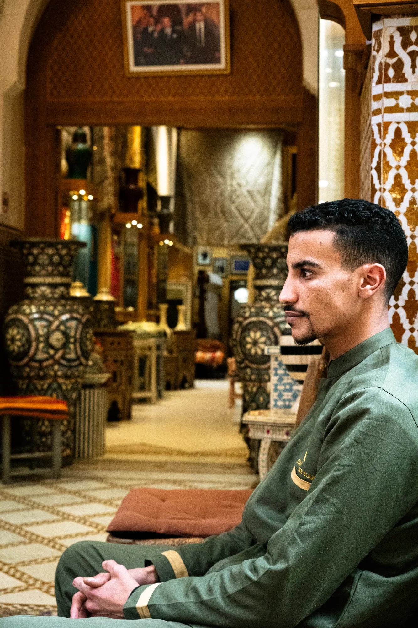 A young man in a green djellaba in profile, a long shop interior behind him