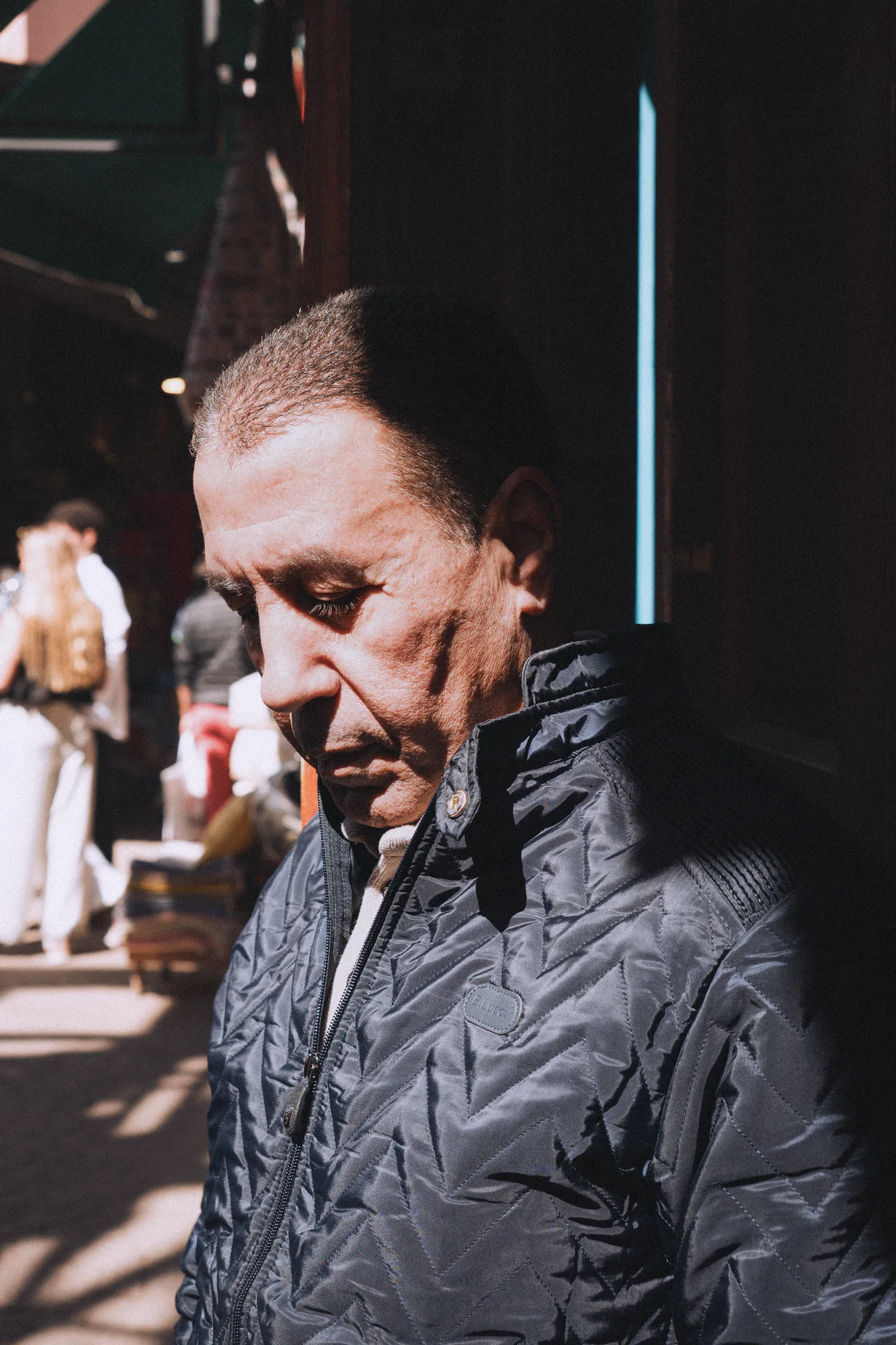 An older man in a quilted jacket, a single band of sunlight across his face in a dark souk