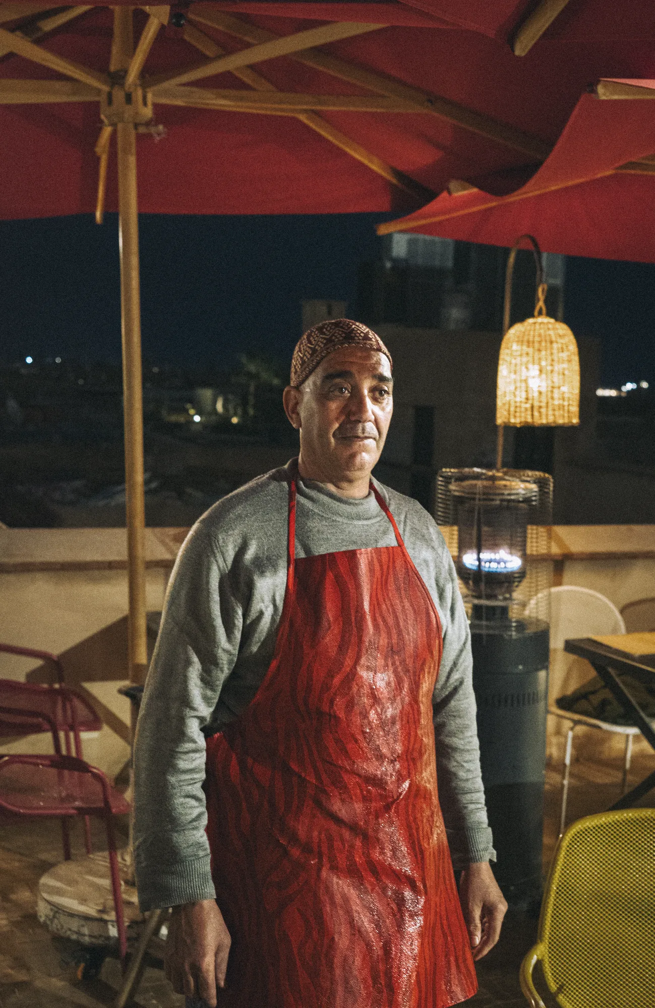 A waiter in a red apron and embroidered cap on a rooftop at night