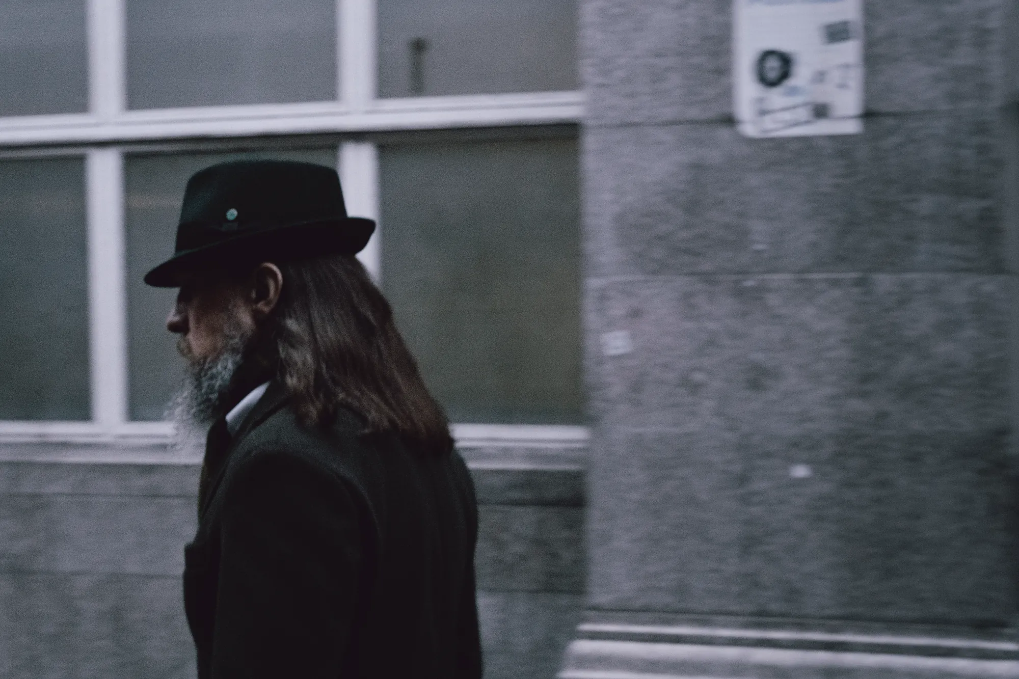 A man in a black hat and long coat walking past a window