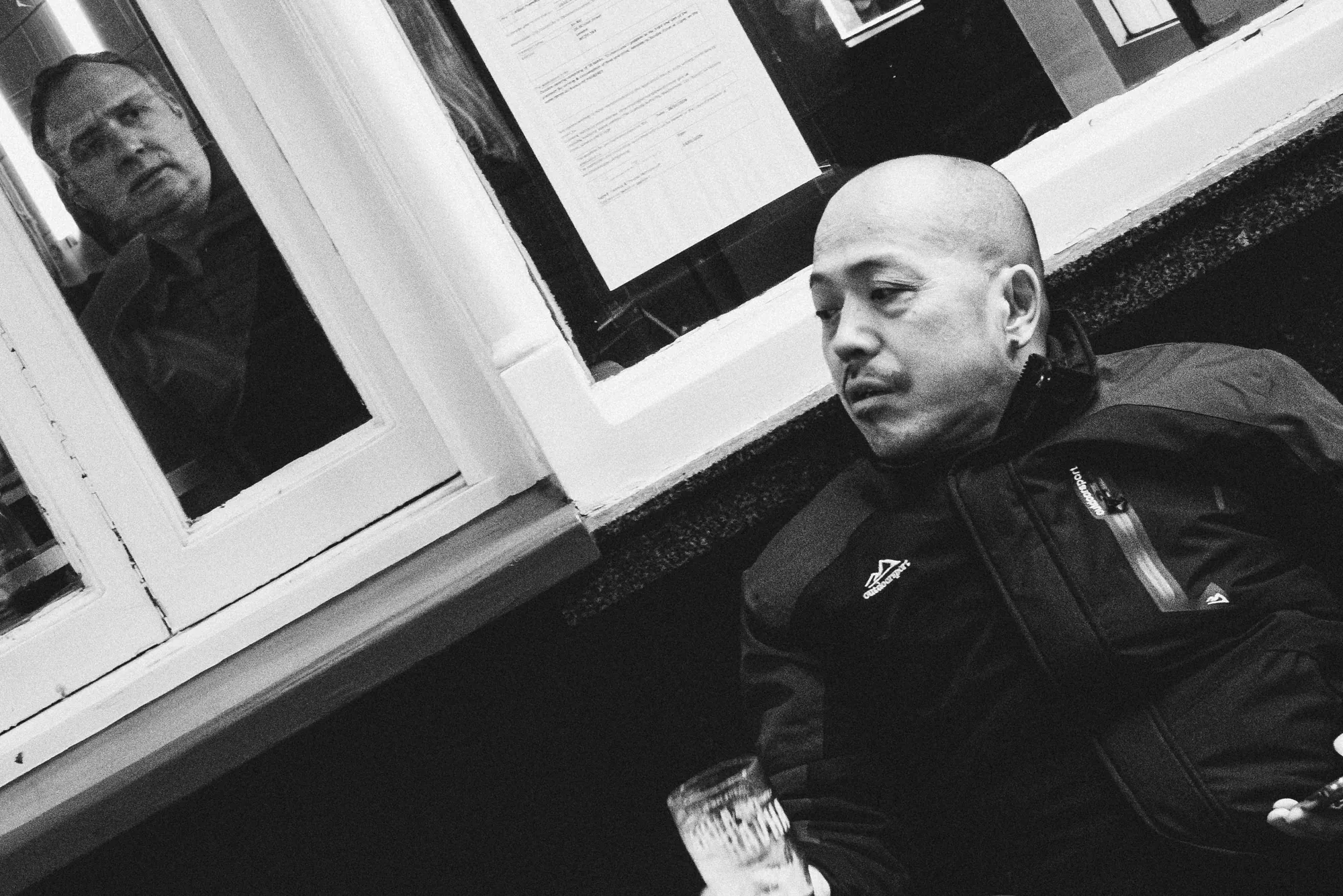 A man with a pint outside a pub while another man looks out through the window behind him