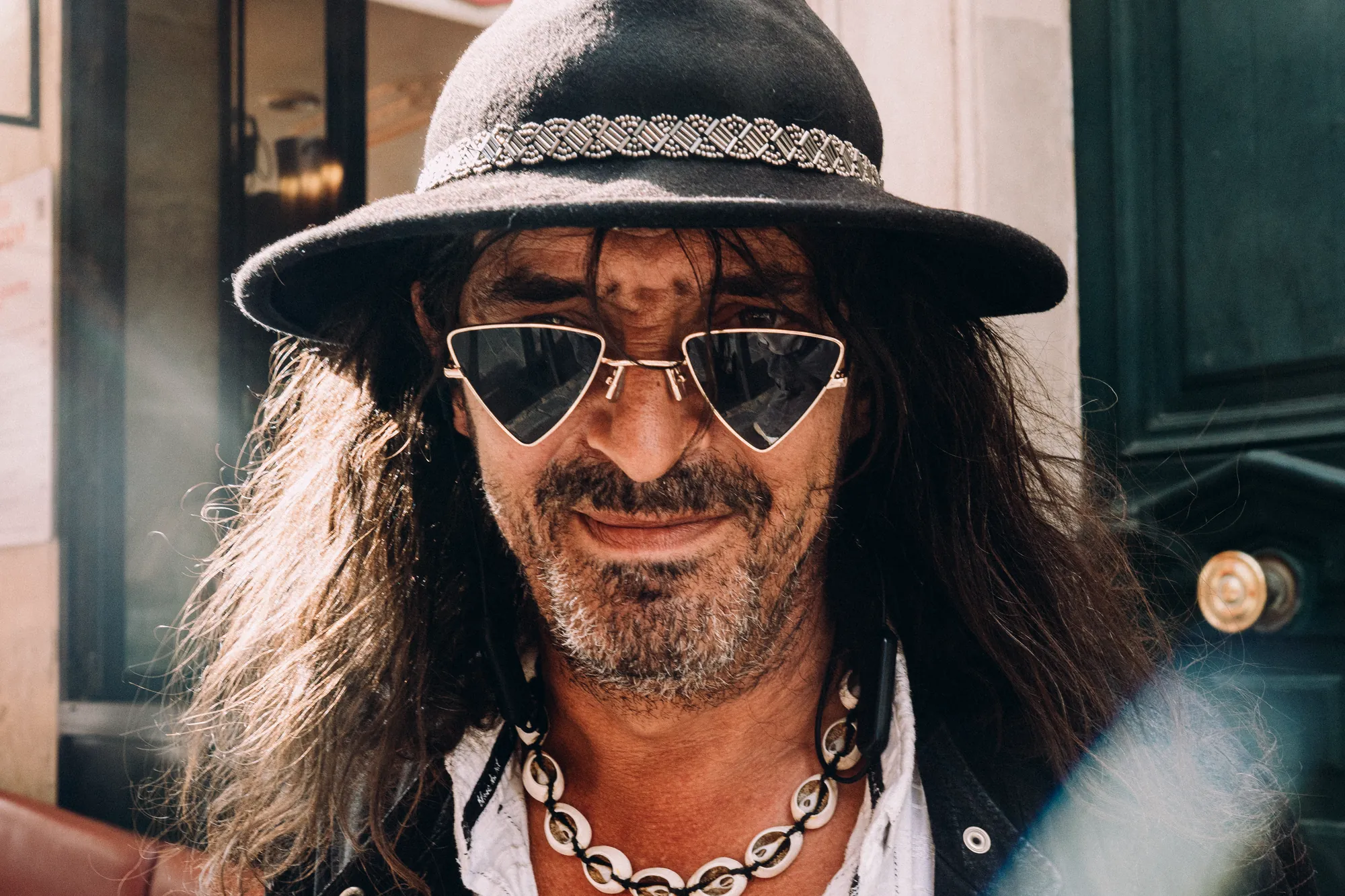 A man with long hair, a black hat and triangular sunglasses, smiling