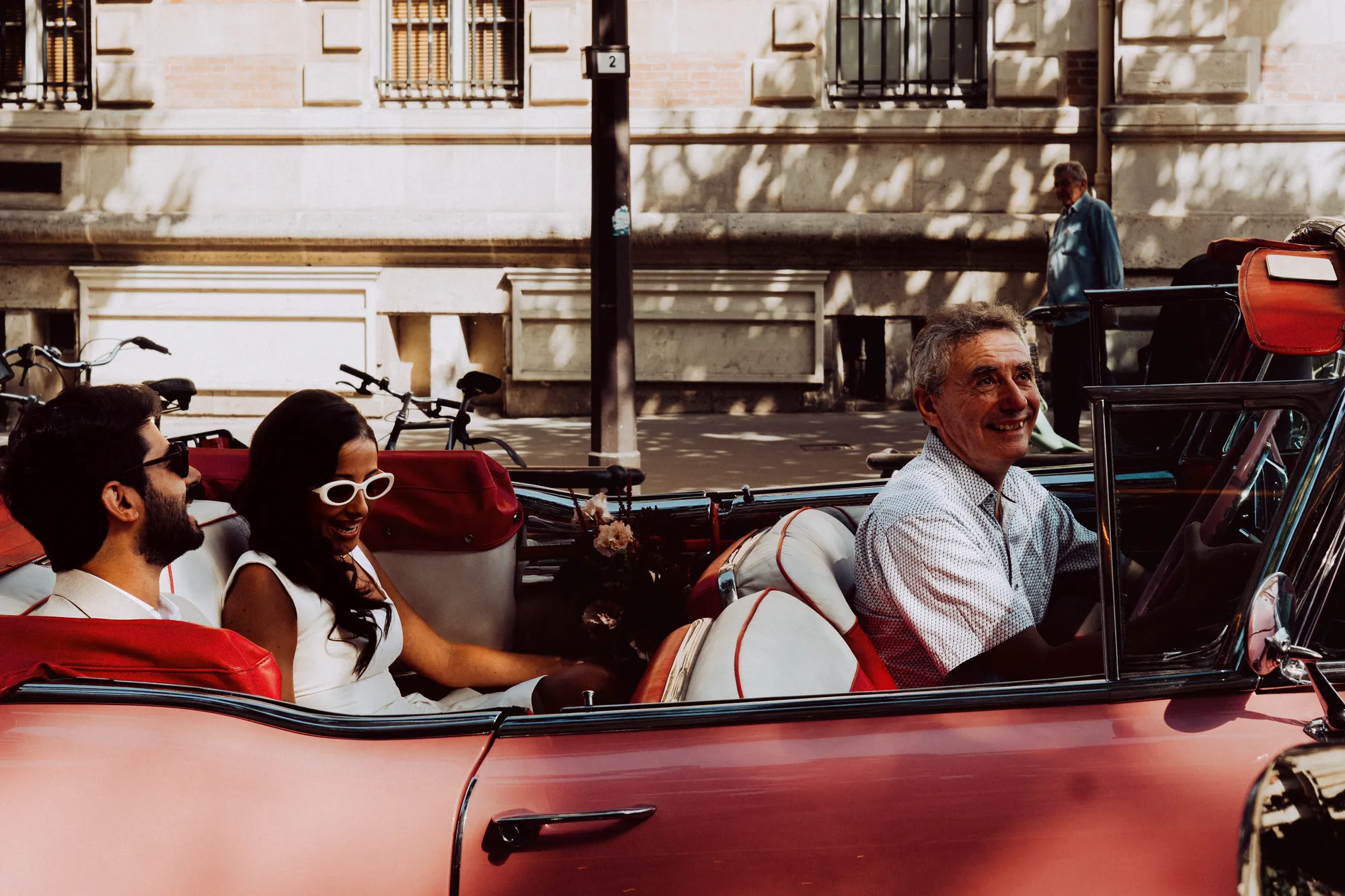 A couple laughing in the back of a pink convertible, an older man turning round from the front seat